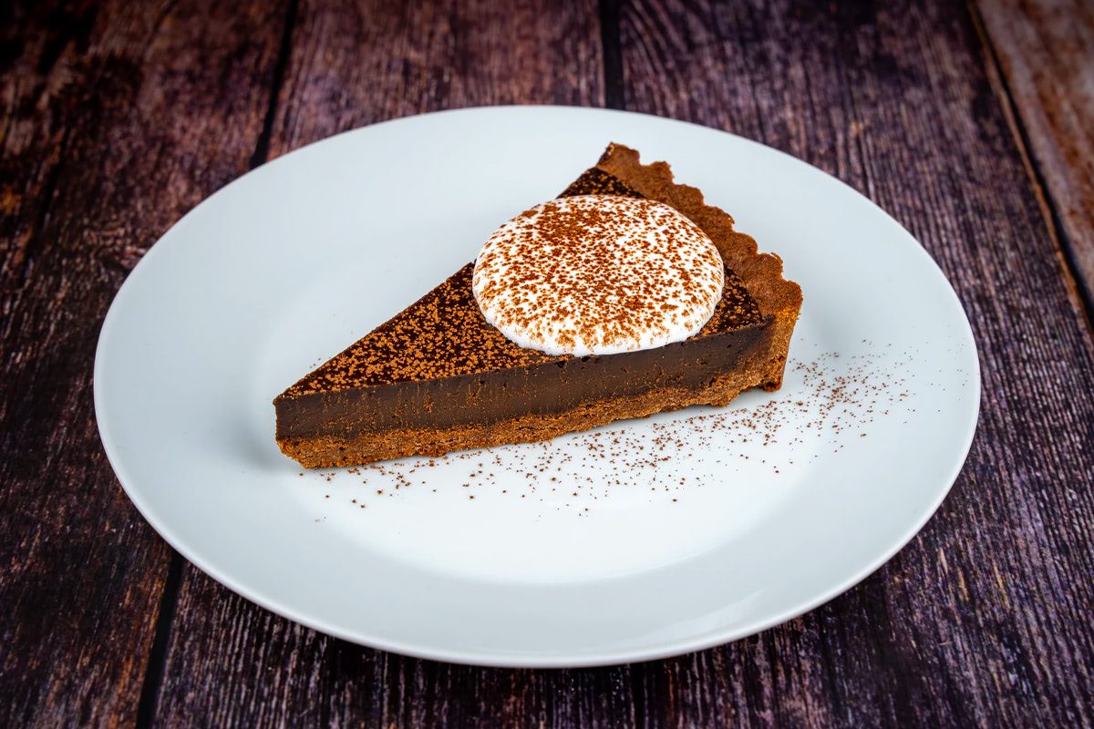 Porción de tarta de chocolate con base de galleta, cubierta de crema batida y espolvoreada con cacao en polvo, servida en un plato blanco sobre una mesa de madera.