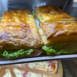 Pasteles de hojaldre dorados rellenos de jamón y lechuga, exhibidos en un mostrador junto a una pizza.