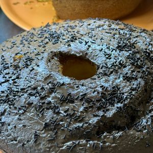 Pastel de chocolate decorado con chispas negras, con un agujero en el centro, acompañado de un bizcocho amarillo en un plato naranja. Ideal para celebraciones y postres.